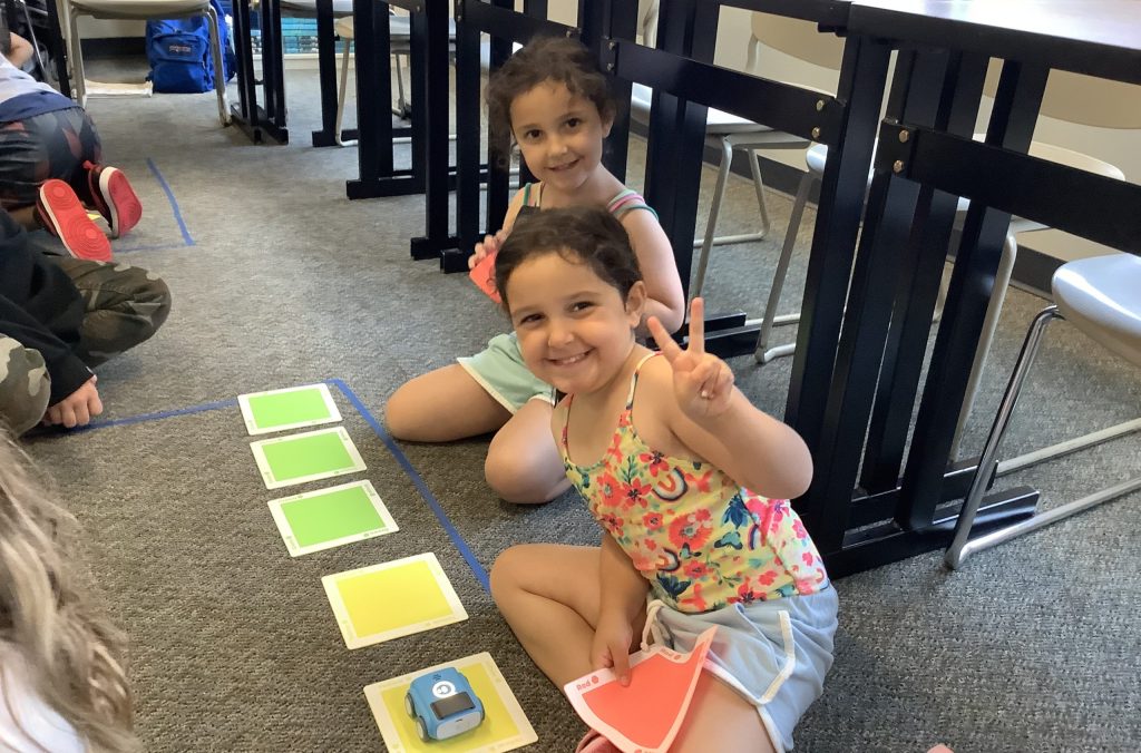 A young girl with brown hair giving a piece sign to a camera in the middle of an camp activity program with a friend